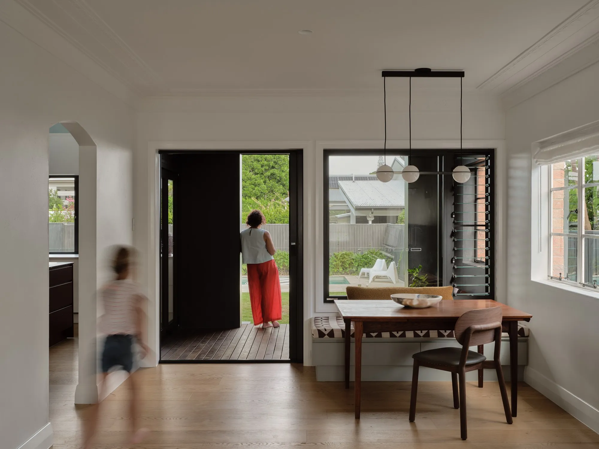 A dining nook with a timber table, patterned banquette and globe pendant, a woman standing in the open back door and a child running past in a blur.