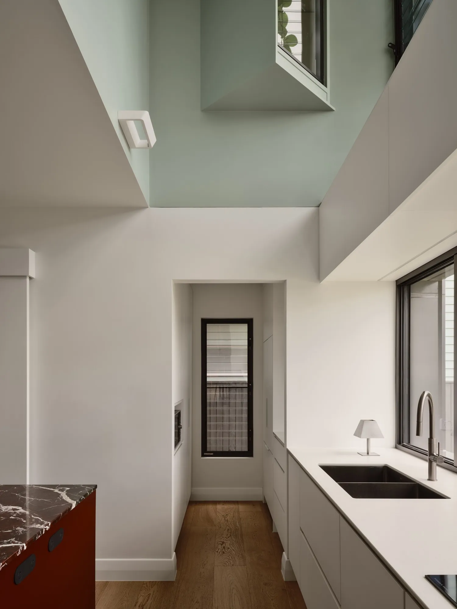 Looking up from the kitchen into a double-height void painted sage, a window box projecting above white joinery, a dark sink and the burgundy marble island.