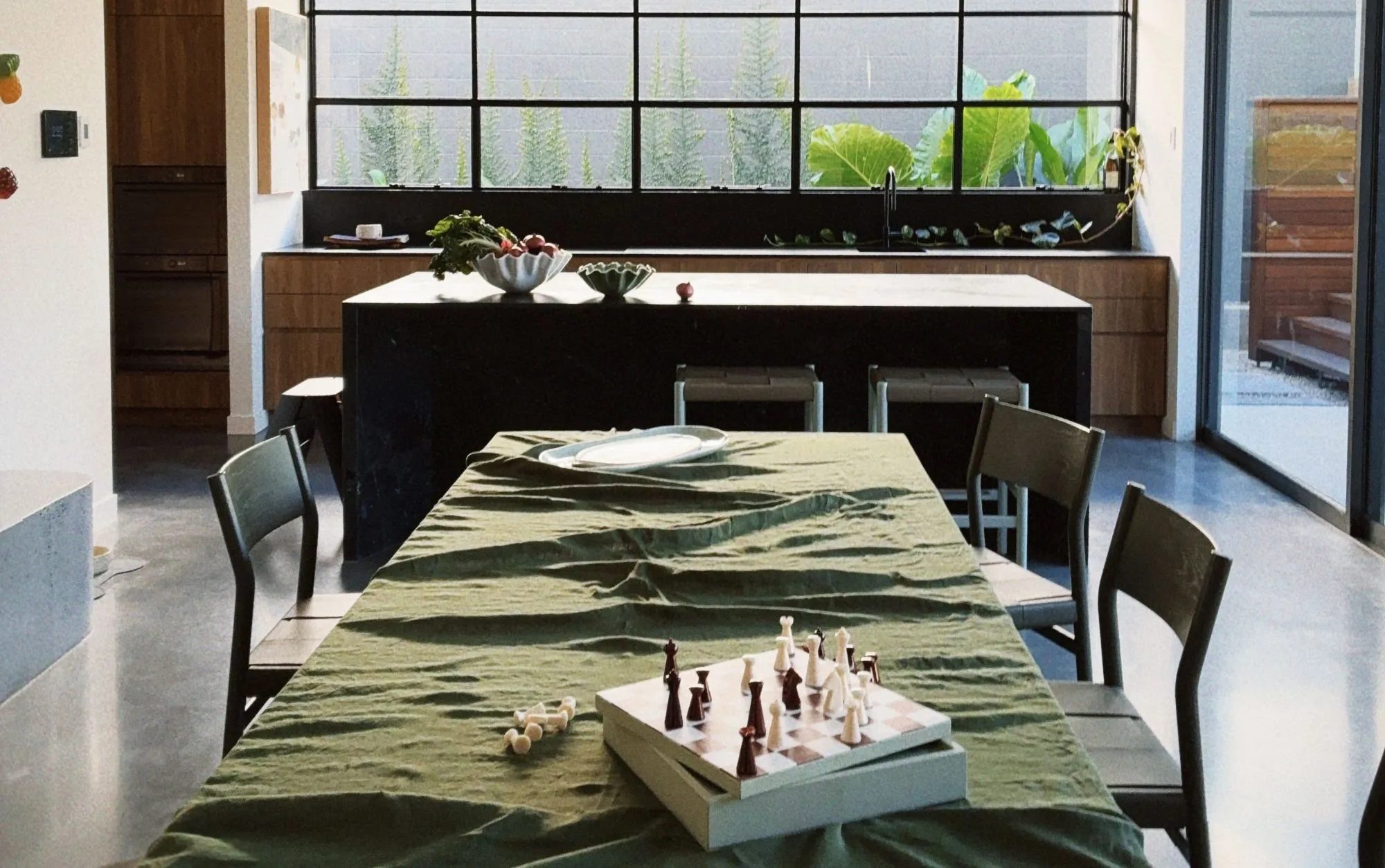 A dining table under a moss green cloth with a chess set mid-game, beyond it a black stone kitchen island, timber joinery and a long window of greenery.