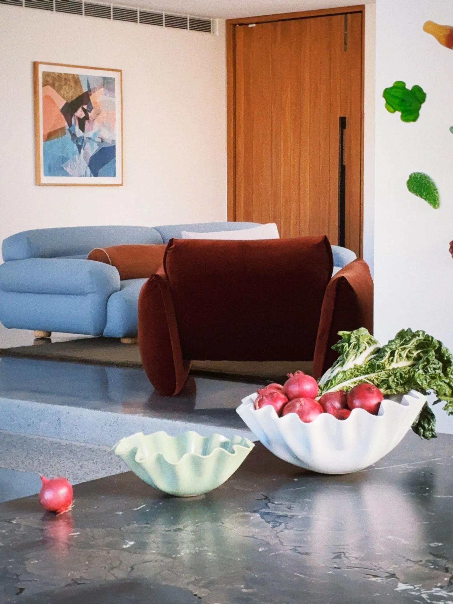 A powder blue sofa with a rust bolster and a deep red velvet armchair on a raised concrete platform, an abstract painting on the wall behind, fluted ceramic bowls of red onions in the foreground.