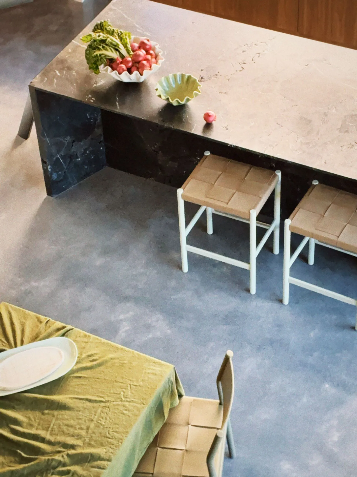 Looking down on a black stone island with a stone top, woven leather stools, and the corner of a velvet-covered dining table on polished concrete.