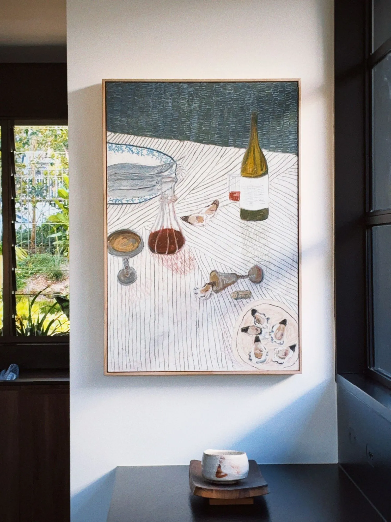 A large framed still life of oysters, wine and a decanter hanging on a white wall in raking afternoon light, a ceramic cup on a timber stand below.