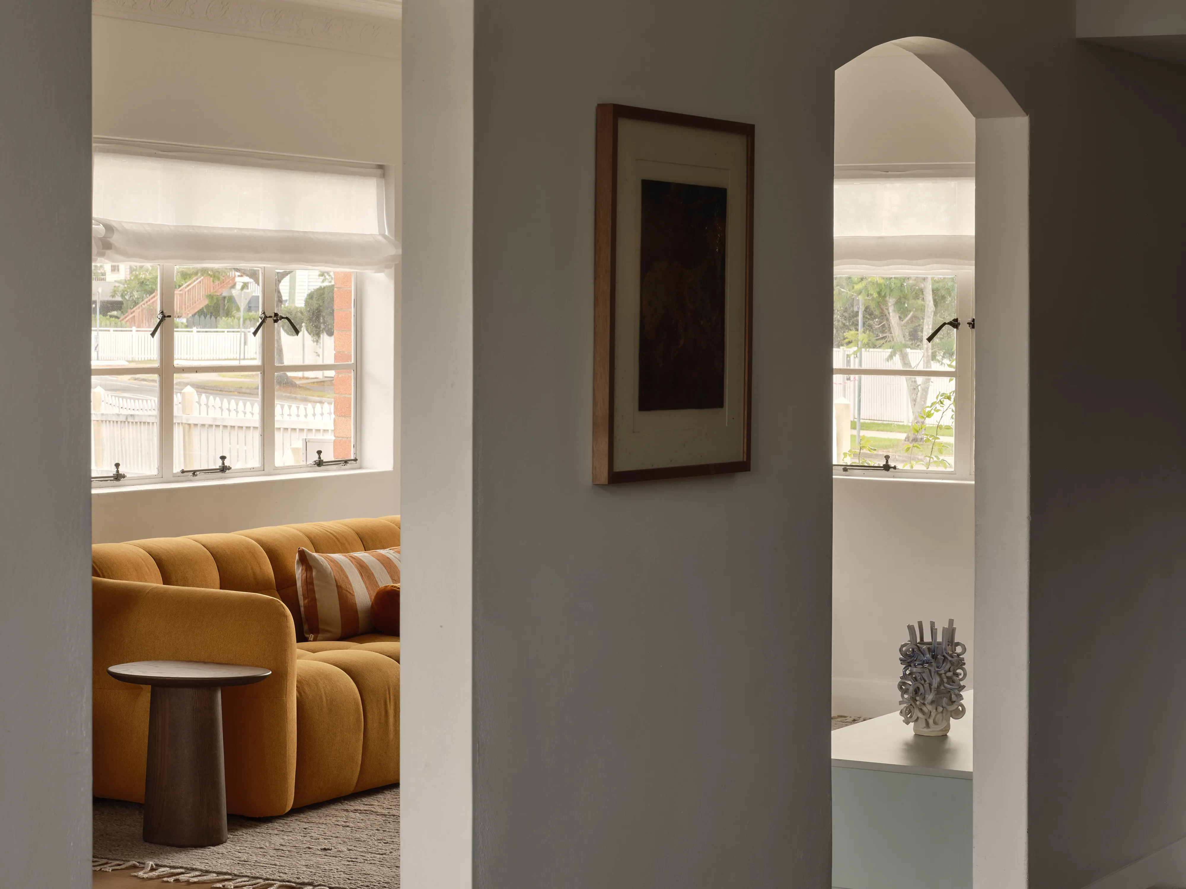 A view between two openings, one to a sitting room with an ochre bouclé sofa and a dark timber side table, the other to an arched alcove holding a pale ceramic vessel.