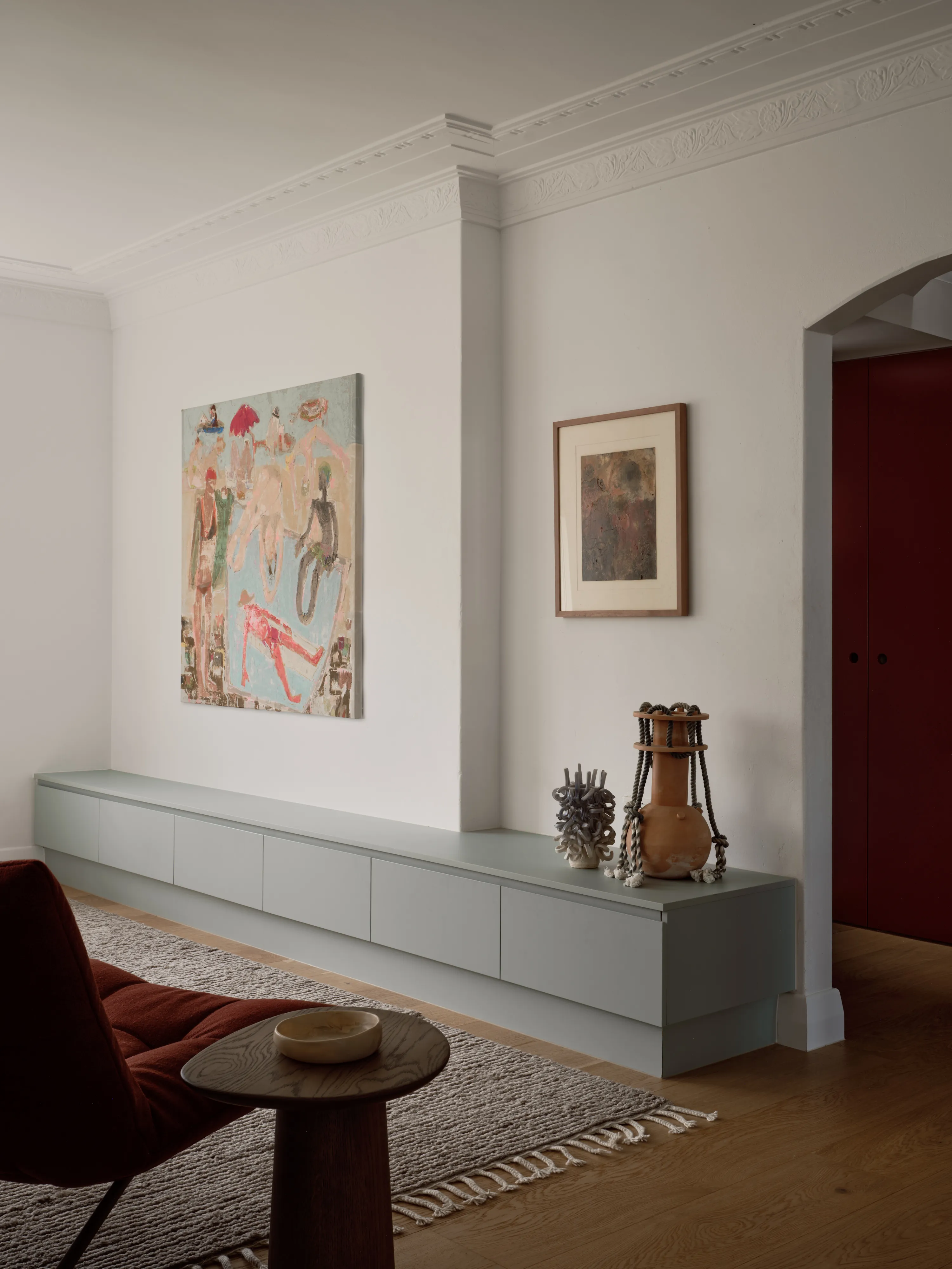 A long sage green sideboard under a large figurative painting, a framed work beside it, a rust bouclé chair and a woven rug in the foreground.