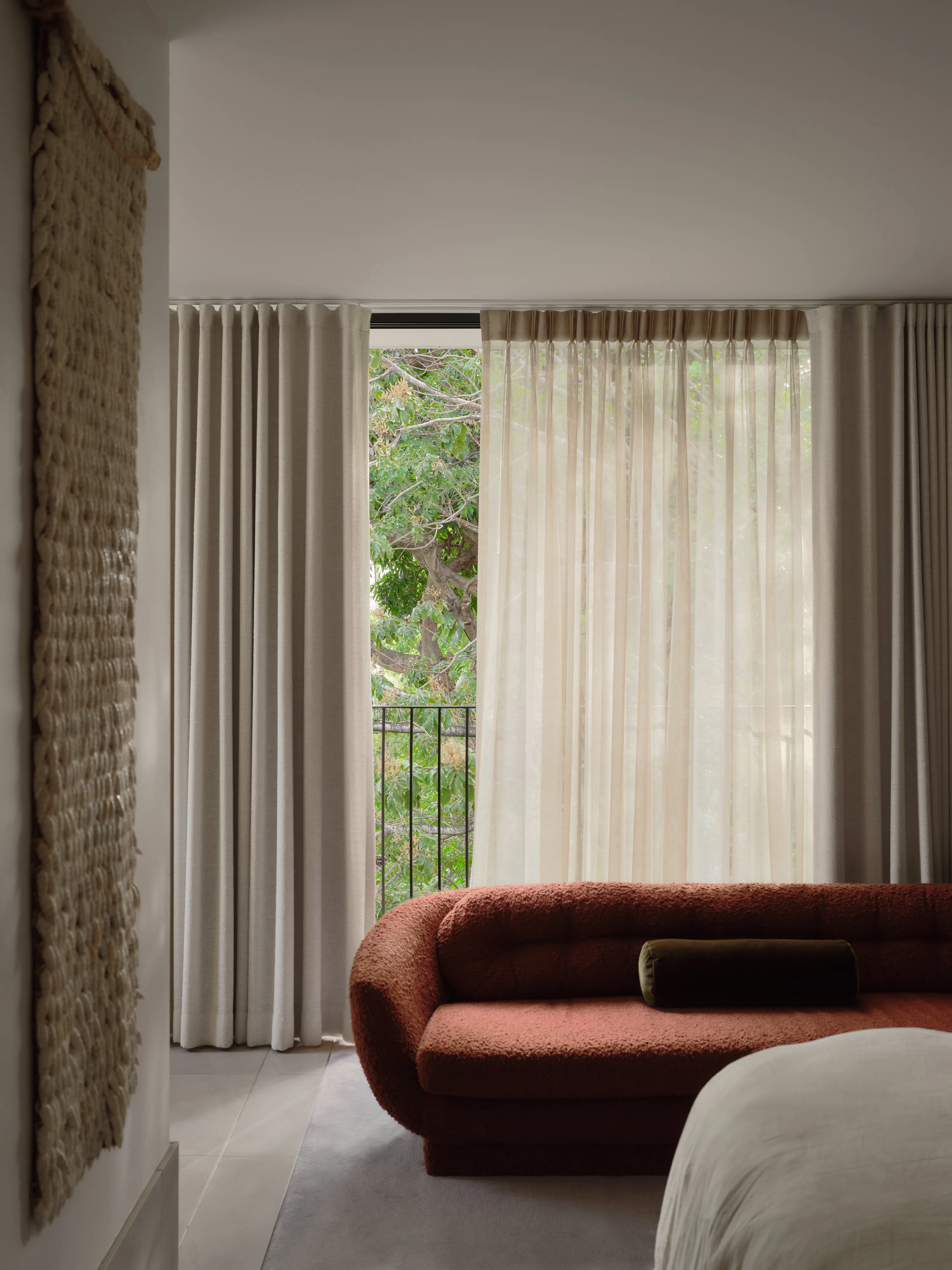 Main bedroom: a curved terracotta bouclé sofa with an olive velvet bolster in front of floor-length linen and sheer curtains, a woven wall hanging at left.