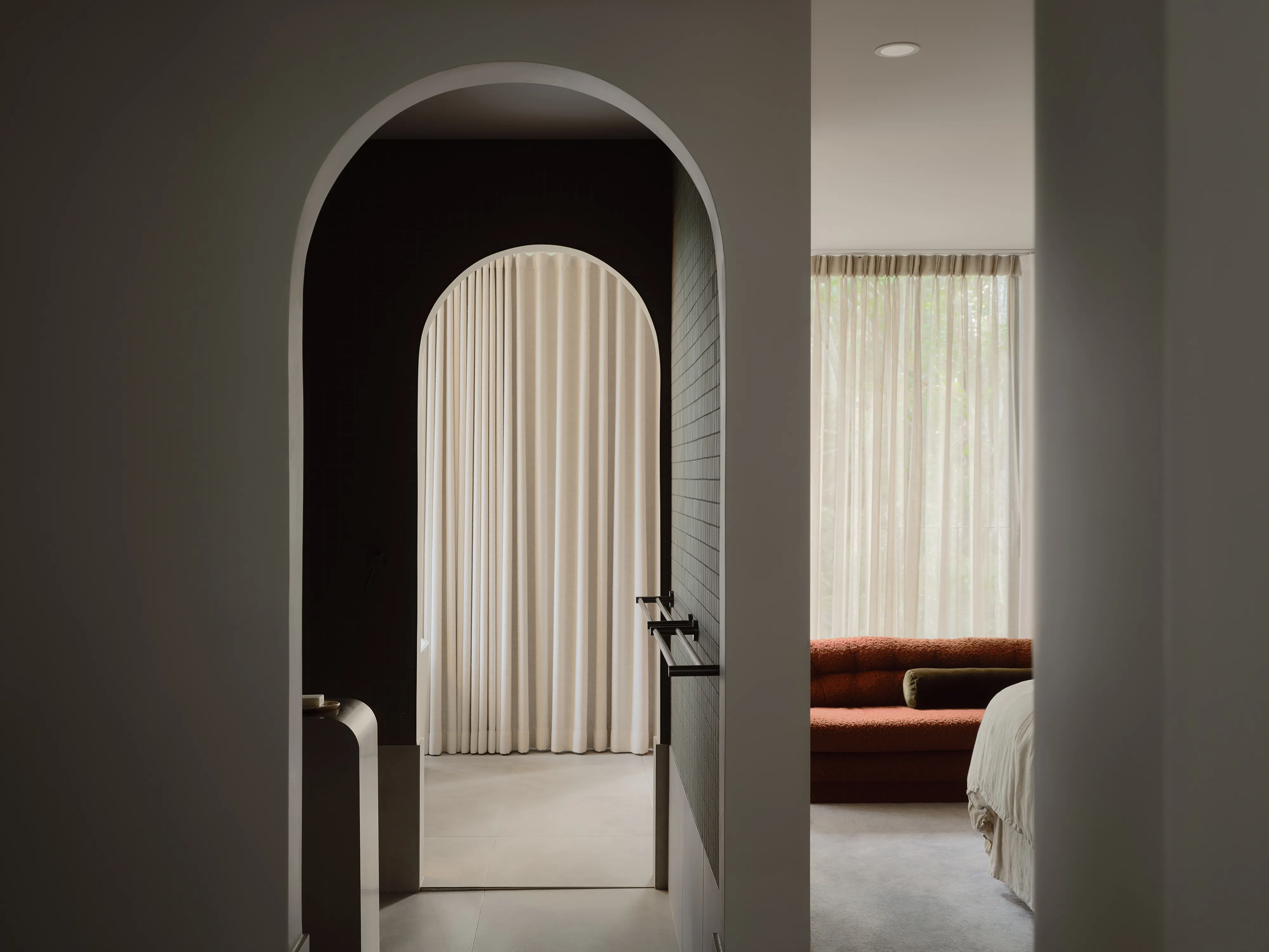 An arched opening into a dark-tiled ensuite, framing an arched linen curtain; beyond, the main bedroom with a terracotta bouclé sofa and sheer curtains.