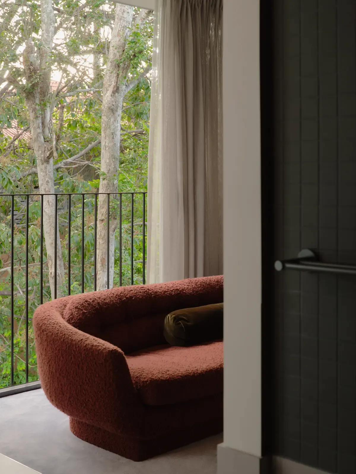 The terracotta sofa beside a balcony balustrade, tree trunks and canopy beyond, a dark tiled wall with a towel rail in the foreground.