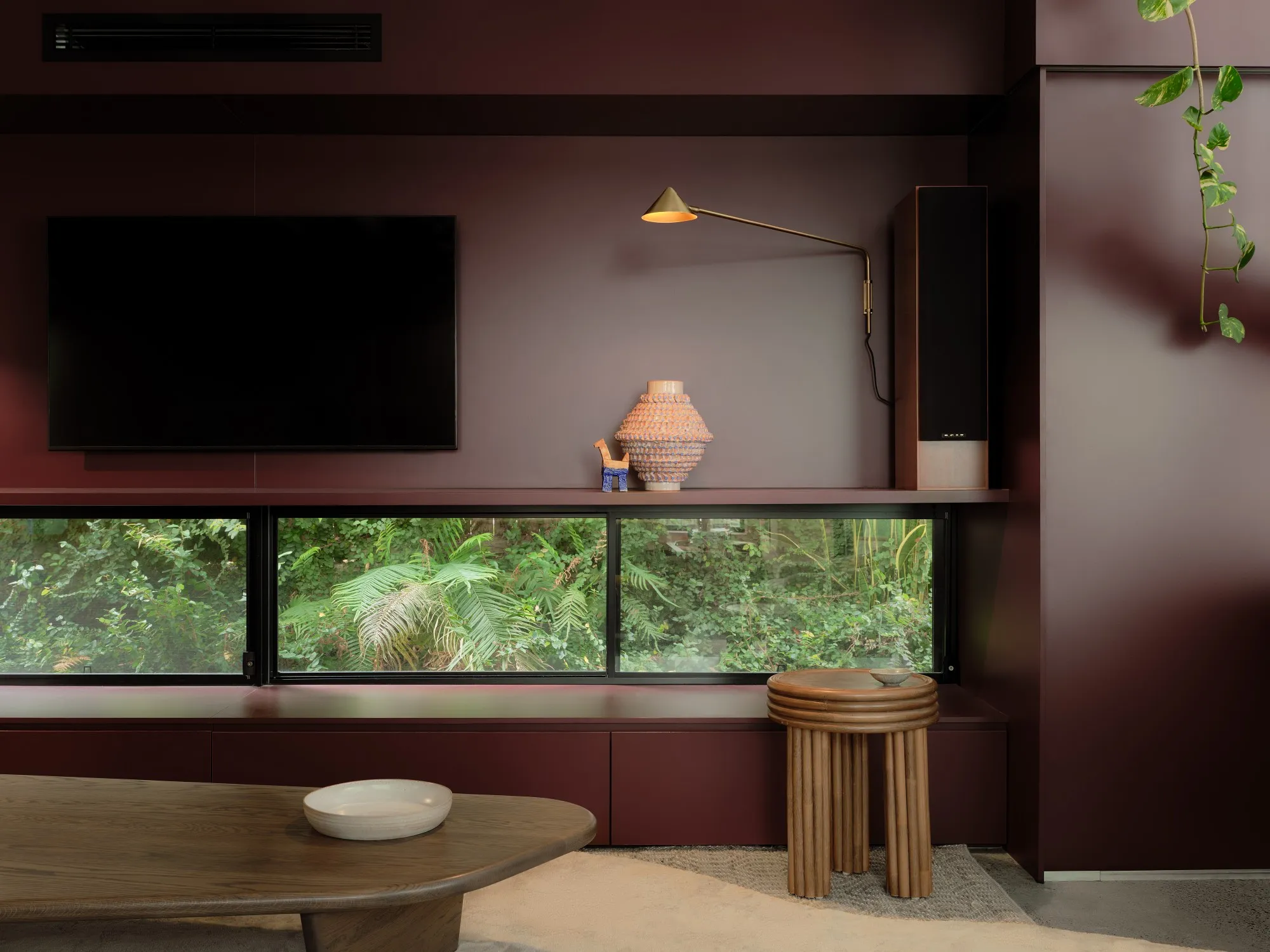 A burgundy media wall with a brass swing-arm lamp and a ceramic vessel on the shelf, a low window onto ferns below, a fluted stool and timber coffee table in front.