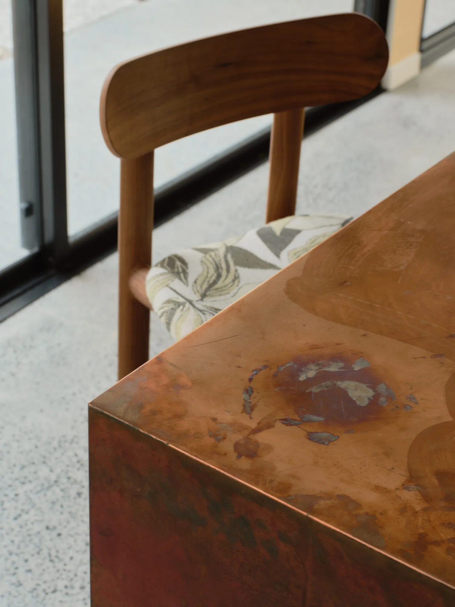 Close detail of a patinated copper table edge beside a walnut chair with a leaf-print seat, polished concrete floor beyond.
