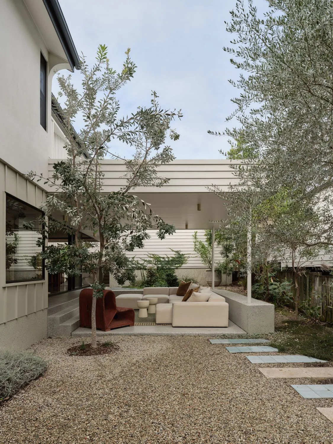 A gravel courtyard with young trees and stepping tiles, a cream modular sofa and a rust woven chair under a white weatherboard canopy.