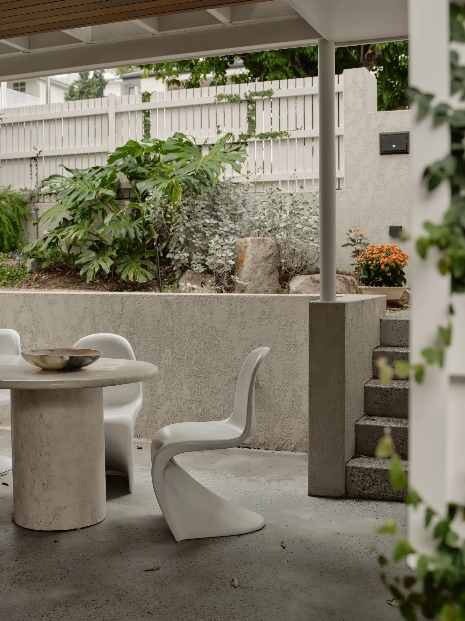 A round travertine table with a marbled bowl and white sculptural chairs under a covered terrace, a rendered wall and planted garden beyond.
